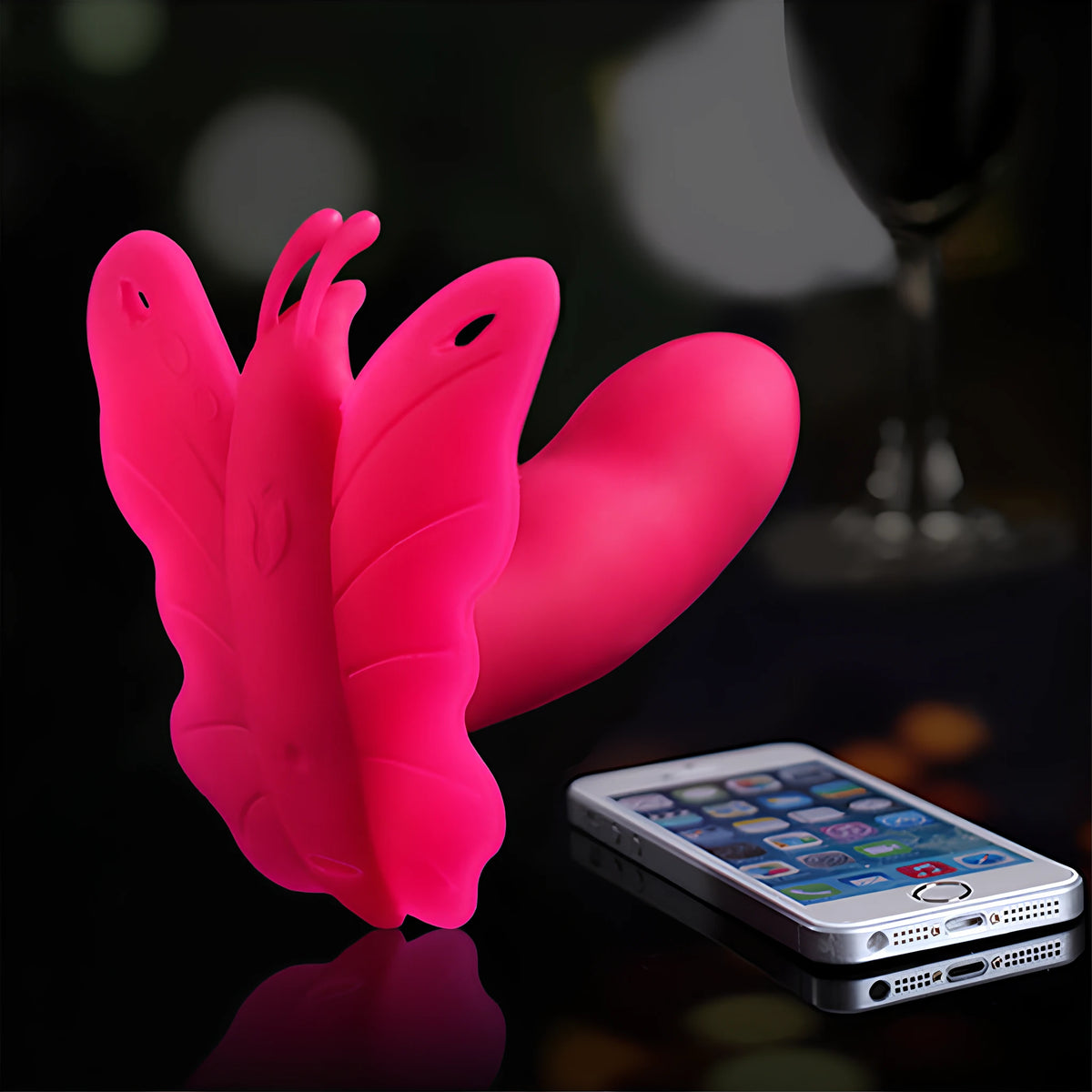 Розовият силиконов вибратор Realov Lydia I Smart Butterfly във форма на пеперуда до смартфон, с който се управлява, на тъмен фон