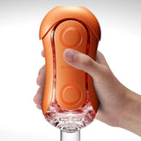 Оранжевият мастурбатор TENGA Flip Orb Sunset Orange, държан в ръка, с два кръгли бутона и прозрачен долен край
